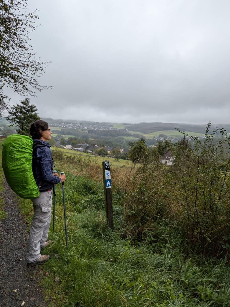 Linda blickt am Druidensteig mit Wanderstöcken und grüner Regenhülle am Rucksack über Dörfer, Wiesen und neblige Hügel.