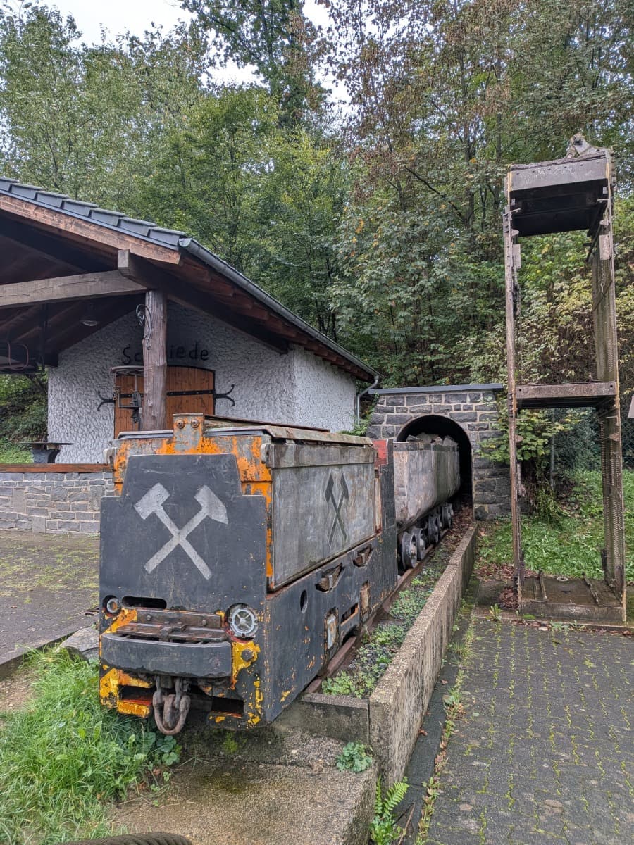 Eine alte Grubenlok mit Loren steht neben der Schmiede des Besucherbergwerks Grube Bindweide im Westerwald vor einem gemauerten Stollenportal.