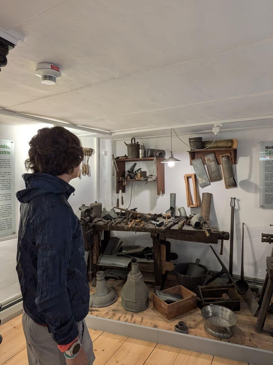Linda betrachtet in eine Ausstellung im Landschaftsmuseum Westerwald mit historischen Werkzeugen und Metallgefäßen auf einer hölzernen Werkbank.