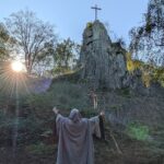 Ein Druide im grauen Kapuzenumhang steht mit ausgebreiteten Armen und einem Holzstab vor dem Druidenstein am Weitwanderweg Druidensteig im Westerwald.