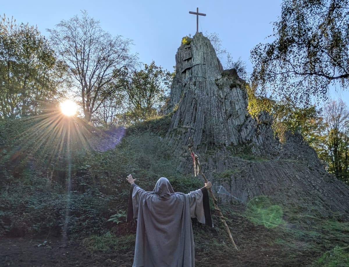 Ein Druide im grauen Kapuzenumhang steht mit ausgebreiteten Armen und einem Holzstab vor dem Druidenstein am Weitwanderweg Druidensteig im Westerwald.