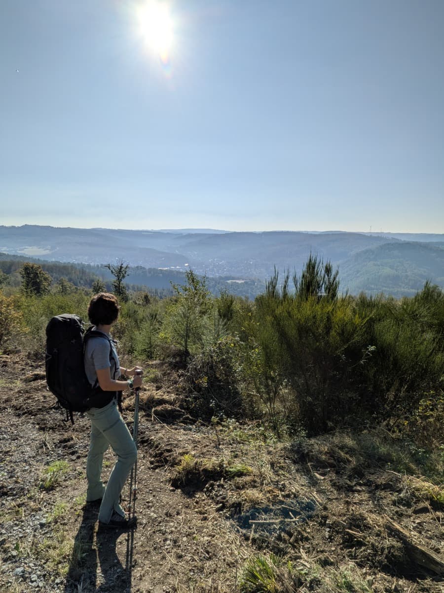Linda blickt während ihrer Mehrtagestour im Westerwald mit Rucksack und Wanderstöcken von einem sonnigen Hang über bewaldete Hügel.