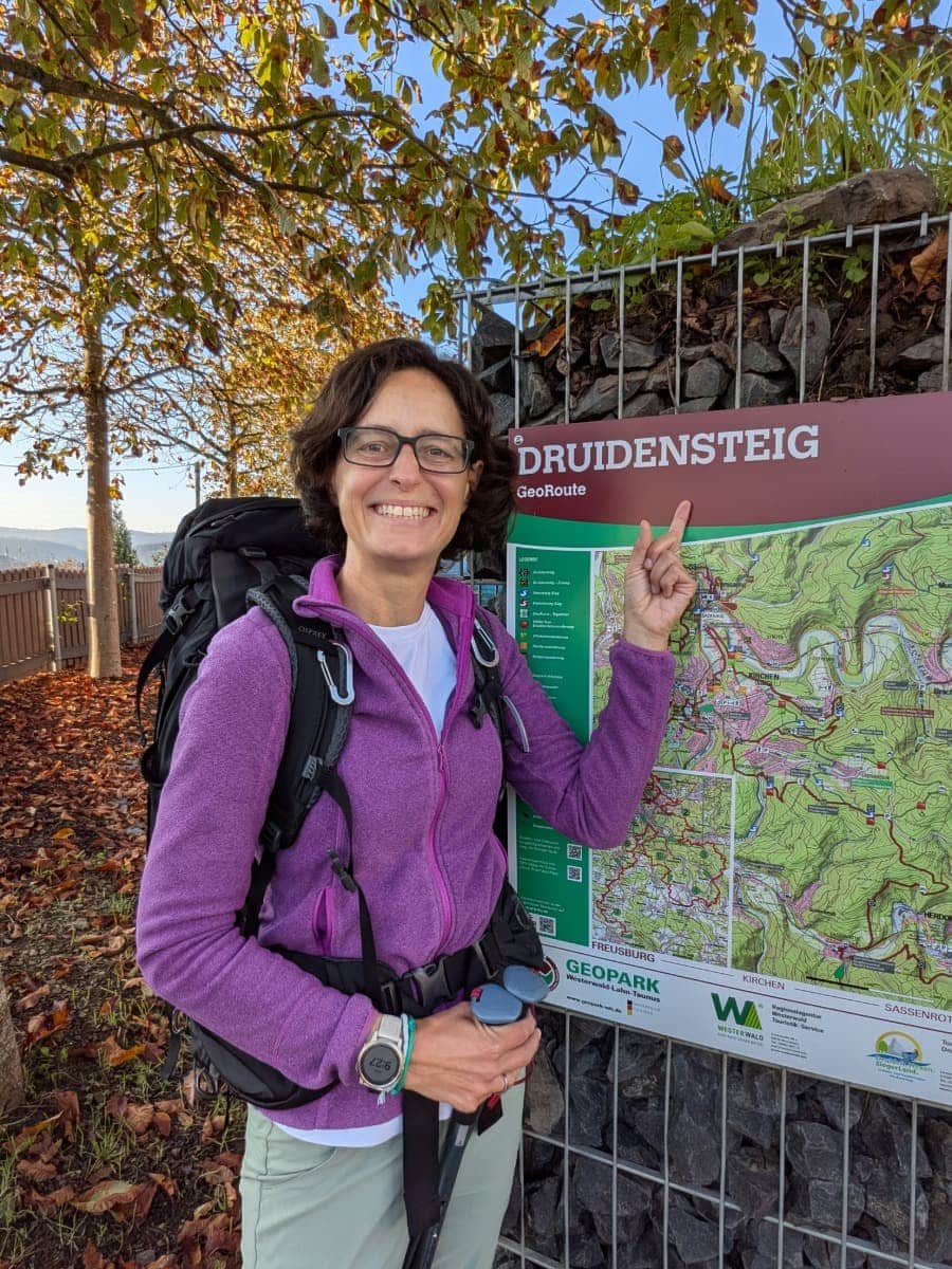 Linda steht mit Wanderrucksack neben einer Wanderkarte und zeigt lächelnd auf die Überschrift „Druidensteig“.