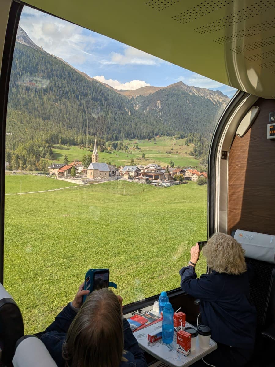 Durch das große Panoramafenster des Bernina Express fotografieren Reisende ein kleines Engadiner Dorf mit Kirche, grünen Wiesen und bewaldeten Berghängen entlang der Bahnstrecke.