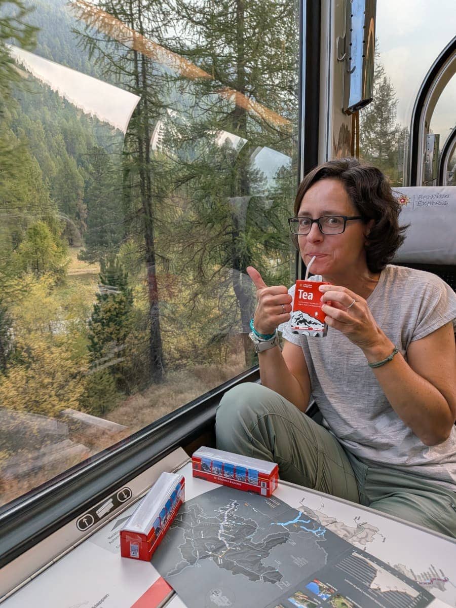 Linda sitzt im Bernina Express am Panoramafenster, trinkt Tee und genießt während der Fahrt den Blick auf die herbstlich gefärbte Berglandschaft.