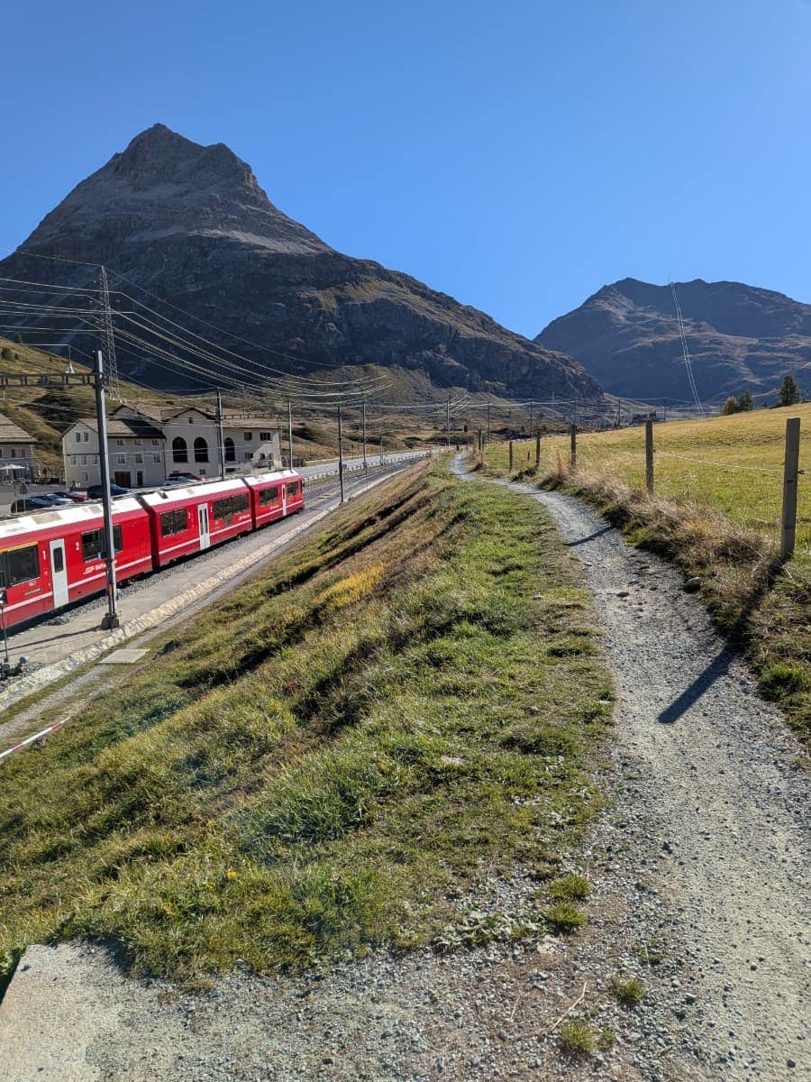 Ein roter Zug der Rhätischen Bahn steht an einer Station an der Berninalinie, während sich daneben der Via Albula/Bernina Wanderweg durch die offene Hochgebirgslandschaft unter markanten Felsgipfeln zieht.