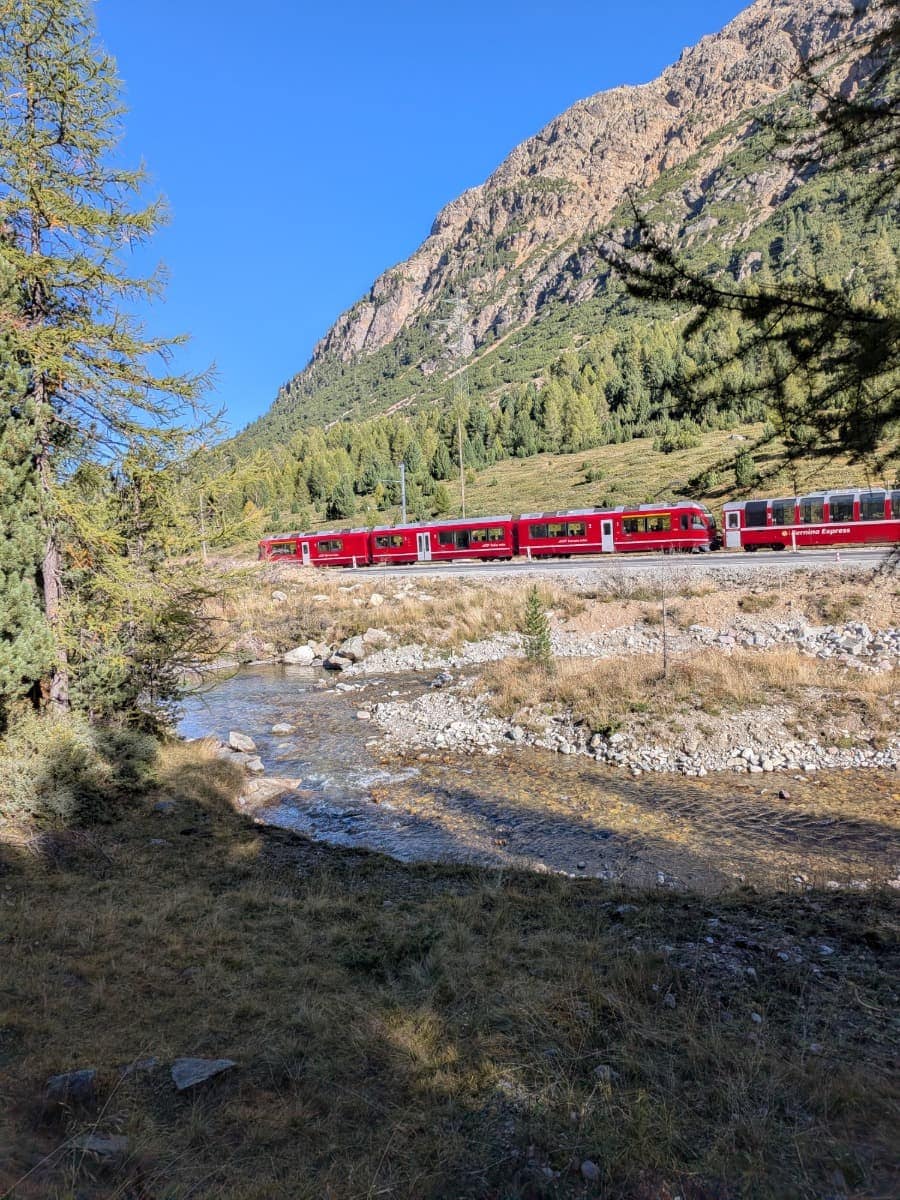 Parallel zum Wanderweg der Via Albula/Bernina fährt ein roter Bernina Express der Rhätischen Bahn durch das herbstliche Hochtal, begleitet von einem klaren Gebirgsbach und steilen Felswänden.