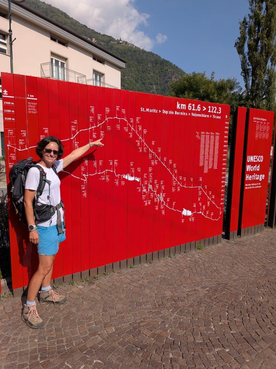Linda steht vor einer roten Übersichtstafel der UNESCO-Welterbestrecke Albula/Bernina und zeigt auf das Höhenprofil der Bahnlinie von St. Moritz über Ospizio Bernina nach Tirano.