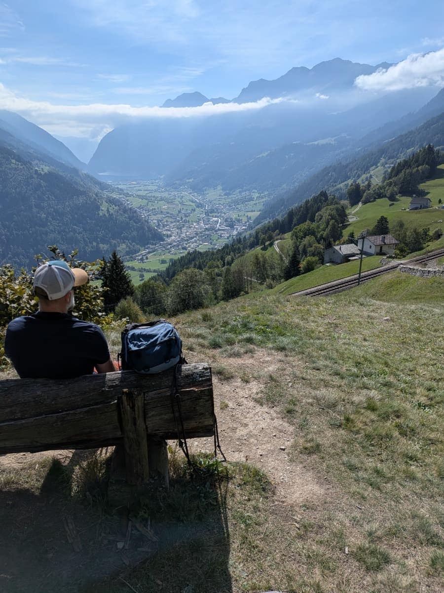 Ein Mann sitzt auf einer Holzbank, vor der sich der Blick über das Val Poschiavo mit dem Ort Poschiavo im Talboden öffnet, während die Strecke der Rhätischen Bahn am grünen Berghang entlangführt.