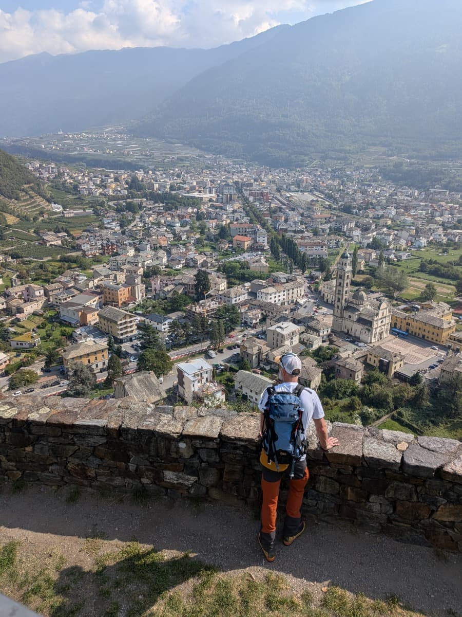 Von einem Aussichtspunkt oberhalb von Tirano blickt ein Wanderer über die dicht bebaute Stadt im Veltlin bis zu den umliegenden bewaldeten Berghängen.