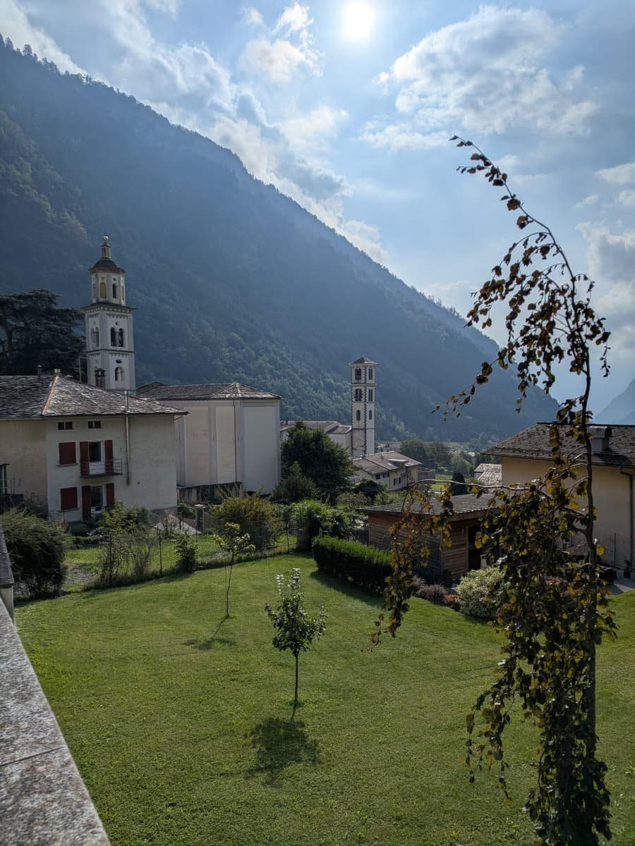 Blick über das Dorf Brusio im Val Poschiavo mit zwei markanten Kirchtürmen, grünen Gärten und steilen, bewaldeten Berghängen im Hintergrund.