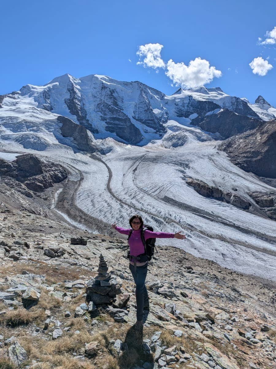 Linda steht mit ausgebreiteten Armen vor dem mächtigen Persgletscher auf der Diavolezza, dahinter ragen die vergletscherten Gipfel der Berninagruppe unter blauem Himmel auf.