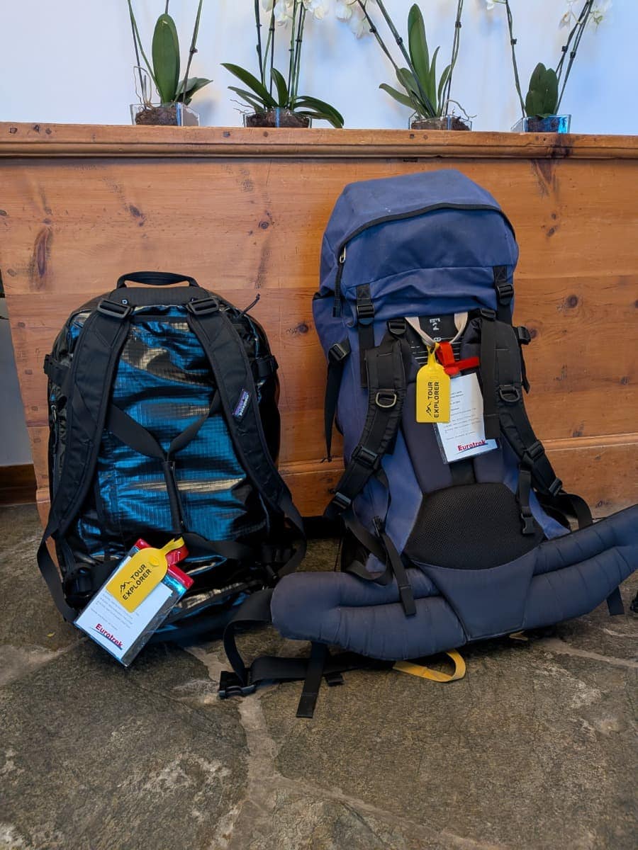 Zwei große Wanderrucksäcke mit bunten Anhänger stehen reisefertig nebeneinander vor einer Holzbank.