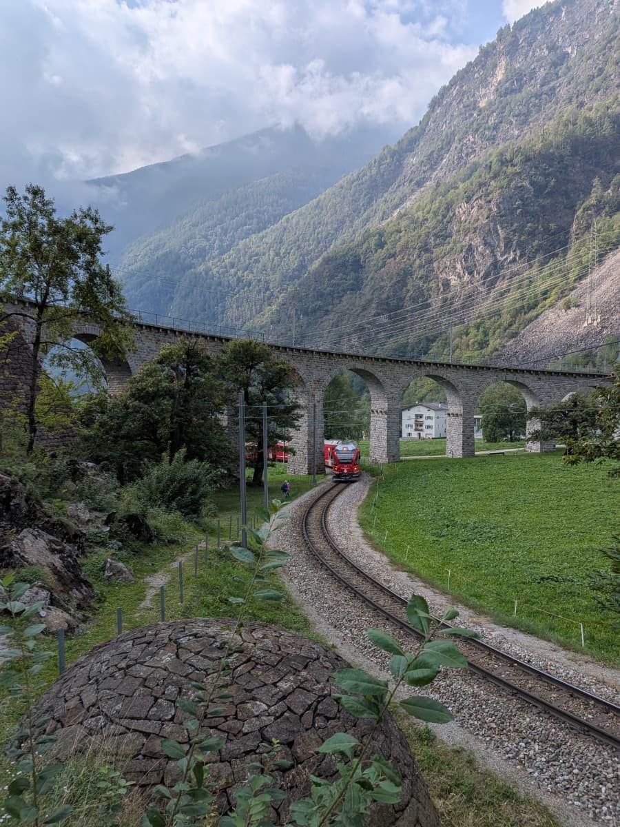 Ein roter Zug der Rhätischen Bahn fährt durch das berühmte Kreisviadukt von Brusio, dessen gemauerte Bögen sich in einer weiten Schleife durch das grüne Tal ziehen. 