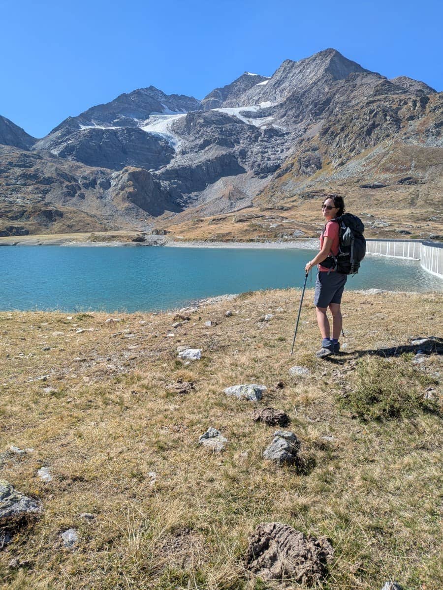 Linda steht mit Rucksack und Wanderstöcken am Lago Bianco, hinter ihr erheben sich die vergletscherten Berge der Berninagruppe, mit Piz Cambrena und dem Cambrenagletscher.