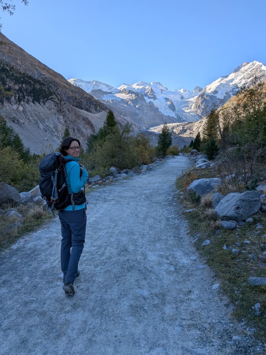 Linda wandert mit großem Rucksack auf einem breiten Schotterweg durch ein herbstliches Alpental, während sich vor ihr der Morteratschgletscher und die schneebedeckten Gipfel der Berninagruppe erheben.