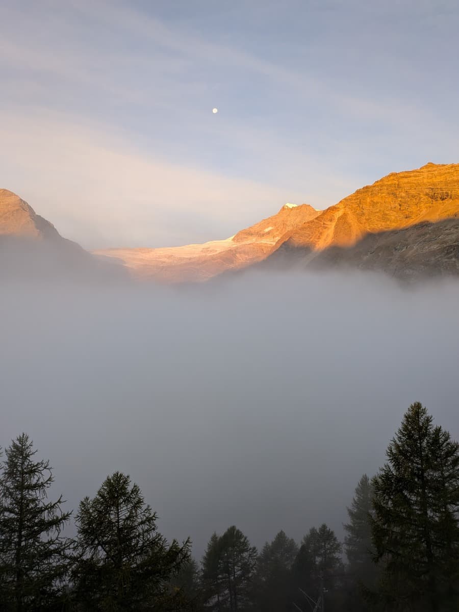Vom Alp Grüm fällt der Blick über den Lagh da Palü zum Palügletscher, dessen Eis und umliegende Felswände im warmen Morgenlicht über einer dichten Nebelschicht leuchten.