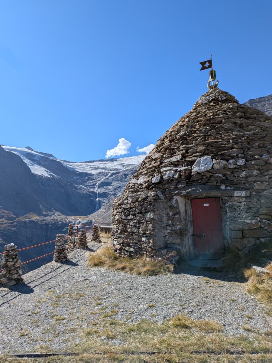Eine kleine Steinhütte mit Schweizer Fahne steht oberhalb des Val Poschiavo, dahinter breiten sich der Palügletscher und die felsigen Flanken der Piz-Palü-Gruppe aus.