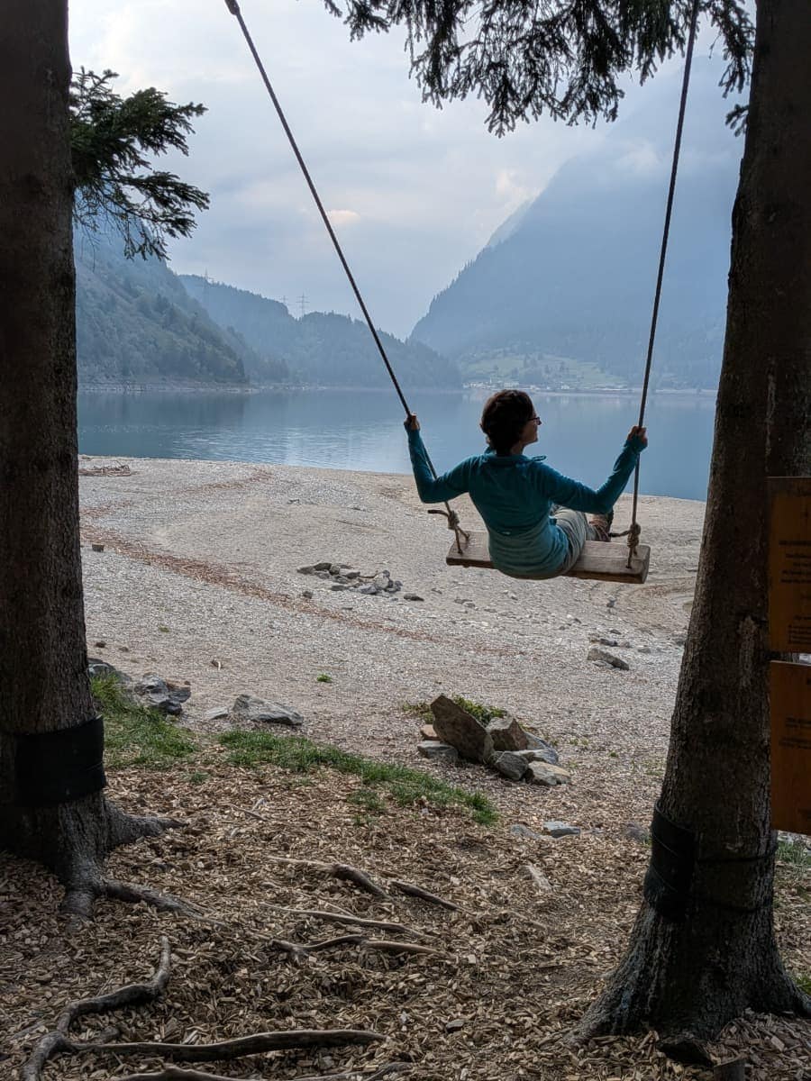 Linda schaukelt zwischen zwei Bäumen am Ufer des Lago di Poschiavo und blickt über den ruhigen See zu den bewaldeten Berghängen des Puschlavs.