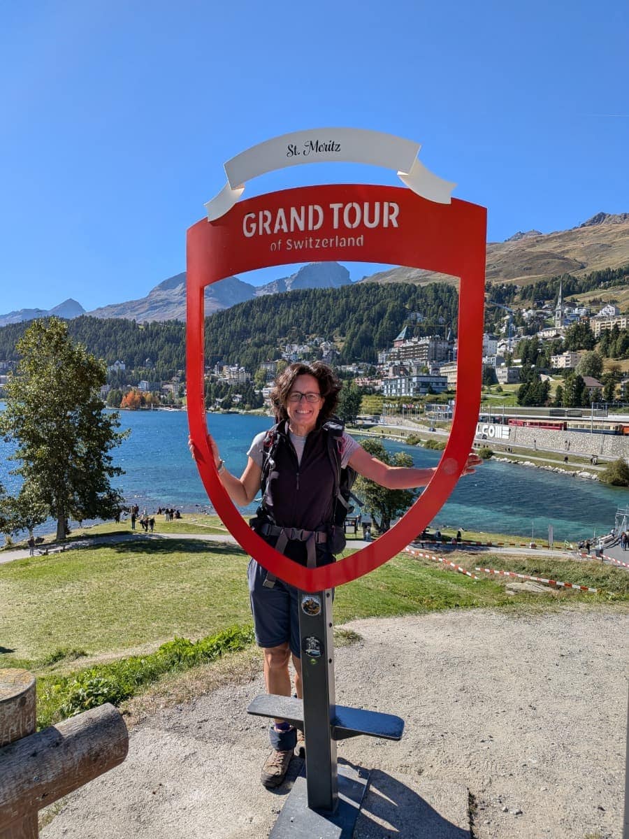 Linda steht lächelnd im roten „Grand Tour of Switzerland“-Fotorahmen oberhalb des St. Moritzersees, mit St. Moritz und den Engadiner Bergen im sonnigen Hintergrund.