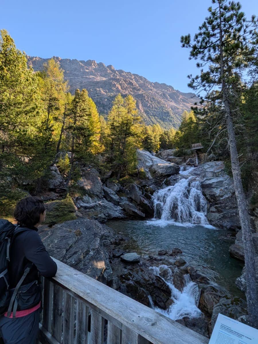 Linda blickt von einer Holzbrücke auf die Ova da Bernina, die auf der Via Albula/Bernina zwischen Felsen und herbstlich gefärbtem Bergwald als kleiner Wasserfall ins Tal rauscht.