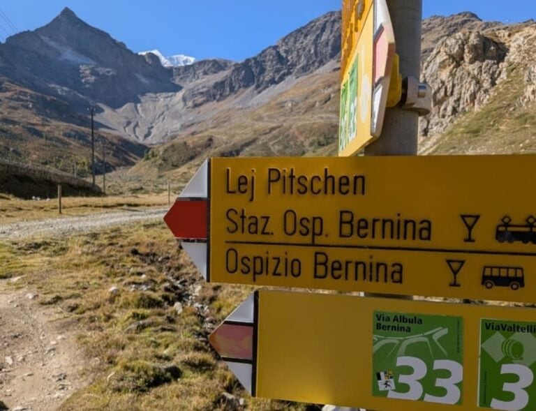 Ein gelber Wanderwegweiser am Berninapass zeigt die Via Albula/Bernina Richtung Lej Pitschen, Staziun Ospizio Bernina und Ospizio Bernina, dahinter steigen karge Hochgebirgshänge zu den Gipfeln der Berninagruppe an.